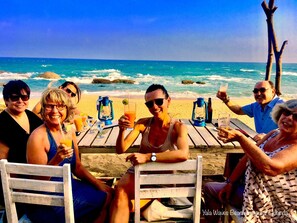 Een groep mensen zit aan een houten tafel op het strand, met de oceaan en rotsen op de achtergrond.
