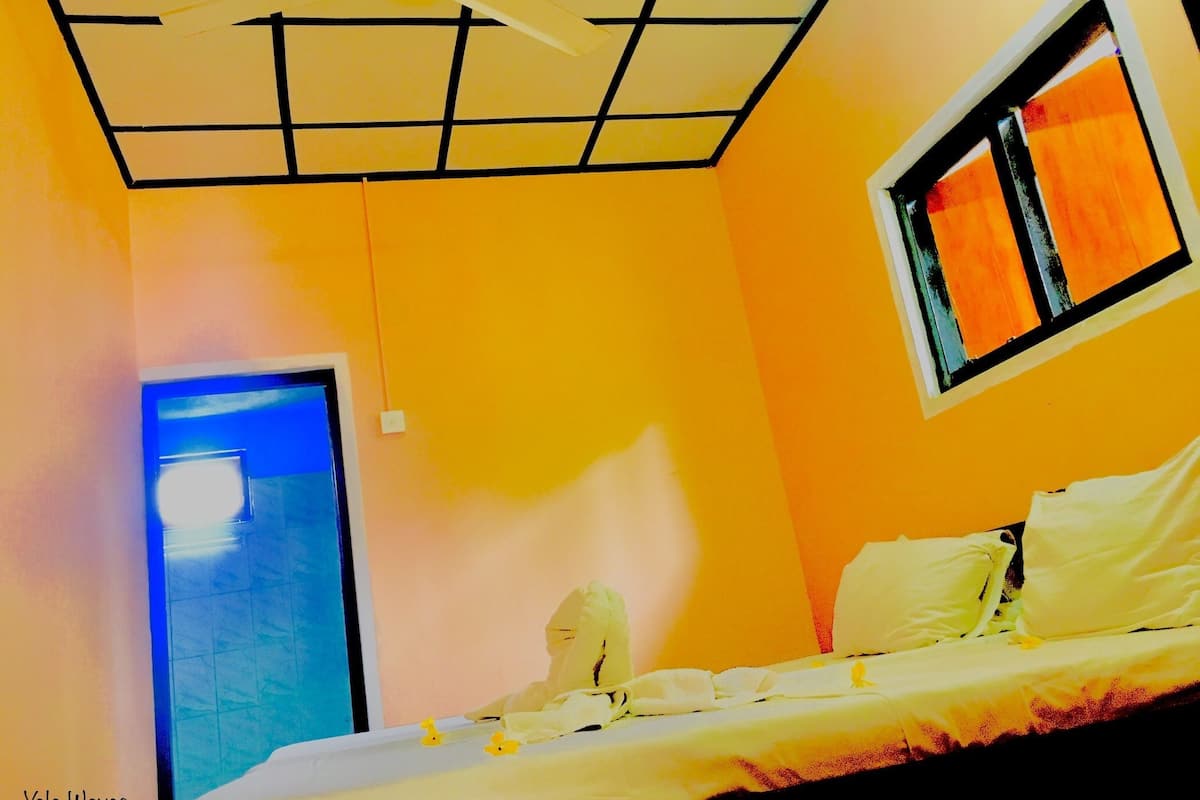 A room with a bed, a ceiling fan, a window with orange curtains, and a door with a light.