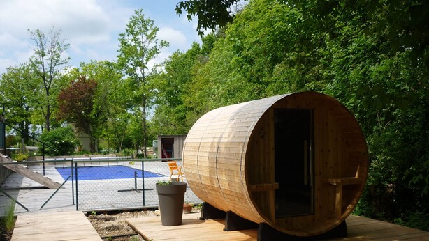 Una estructura de sauna de madera con puerta, un pequeño espacio de estar al aire libre y una piscina al fondo.