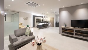 A modern living room with a grey sofa, a coffee table with a vase of flowers, a flat-screen TV on a wooden stand, and a kitchen area in the background. - Castello Hotel (Patras)