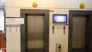 An elevator area with two elevators, each marked with a number sign above. There is a trash bin in front of the elevators, and a digital display screen mounted on the wall between them. - Huaye Hotel (Zhuhai)