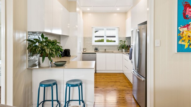 A modern kitchen with white cabinets, a stainless steel refrigerator, a dishwasher, and a window with a view of greenery. There are two blue stools and a potted plant on the counter. - Keeler Cottage (East Toowoomba)