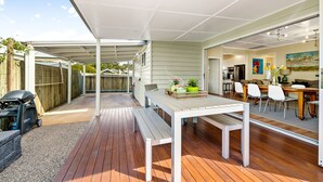 An outdoor patio area with a wooden deck, a white table, and a bench. - Keeler Cottage (East Toowoomba)