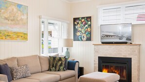 A living room with a beige sofa, a blue ottoman, a fireplace, a television, and artwork on the walls. - Keeler Cottage (East Toowoomba)