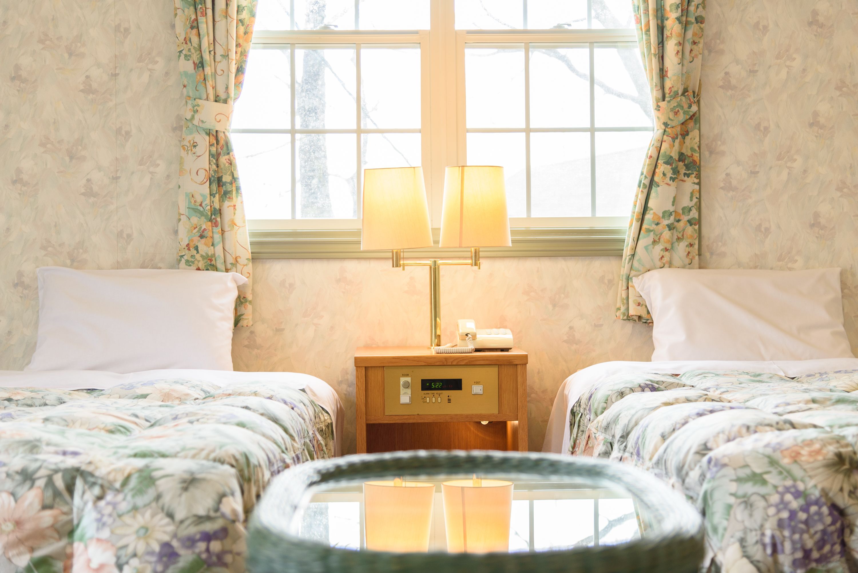A hotel room with two beds, floral bedding, a bedside table with a lamp, and a window with curtains.