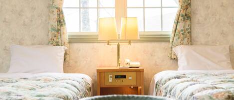 A hotel room with two beds, floral bedding, a bedside table with a lamp, and a window with curtains.