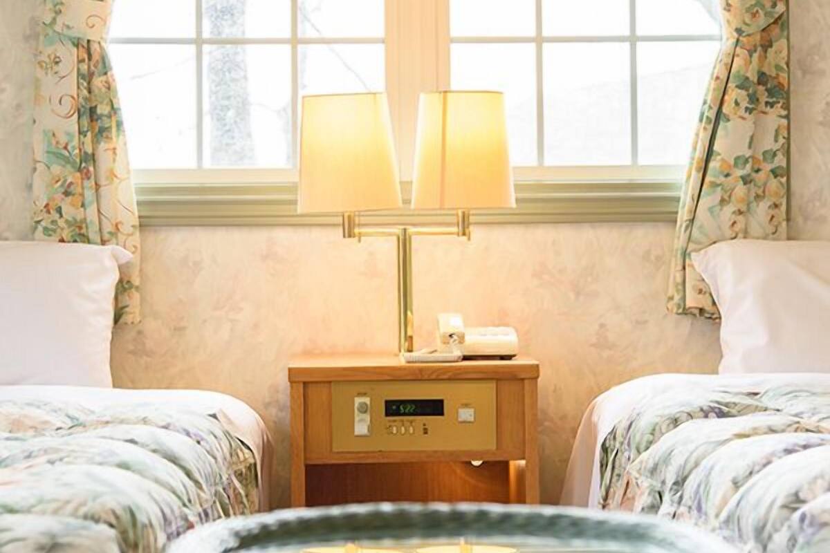Habitación de hotel con dos camas, una mesita de noche con lámpara, un reloj y una ventana con cortinas.