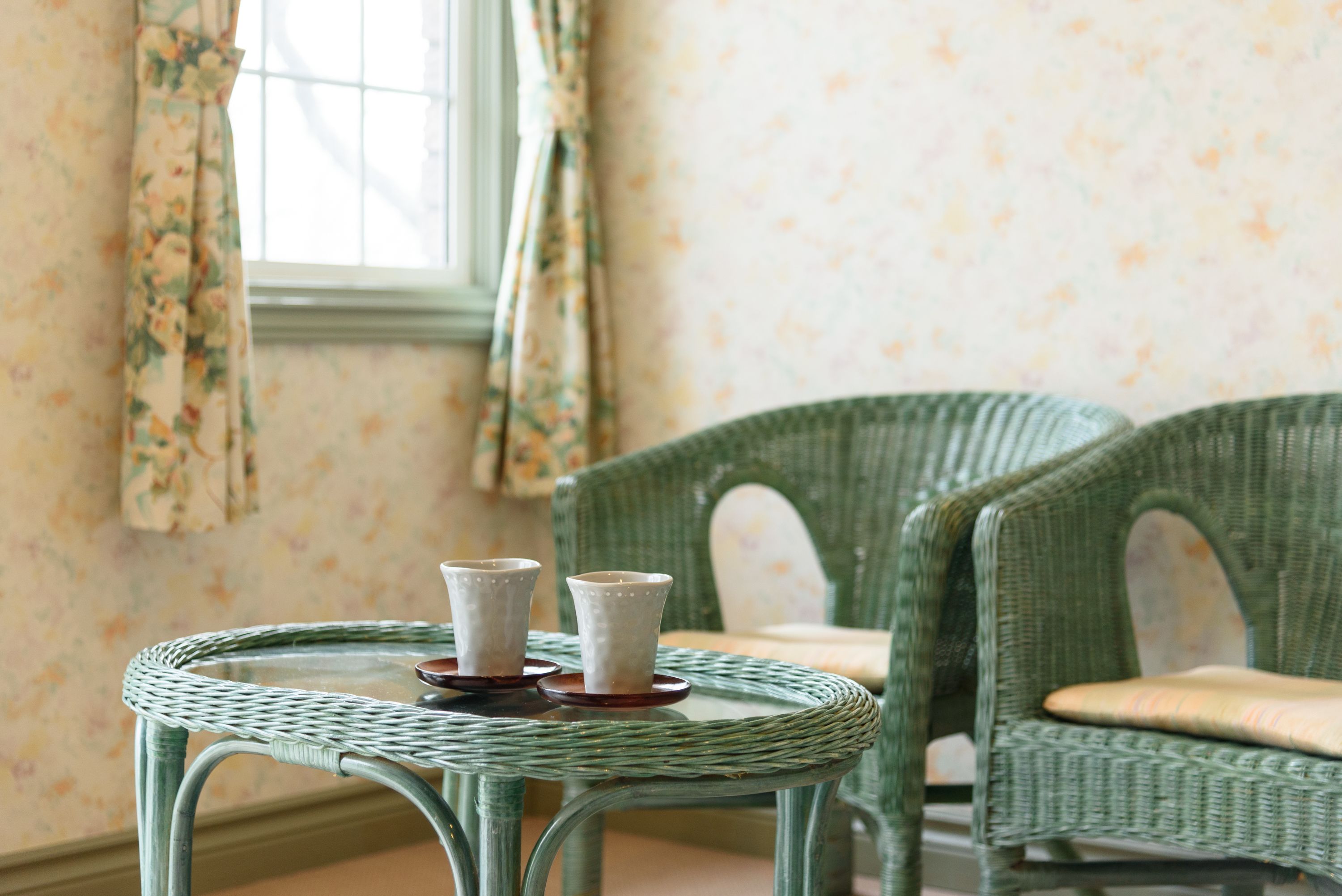 A room with a round wicker table with two white cups on coasters, a green wicker chair, and floral wallpaper.