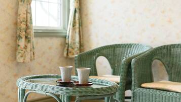 A room with a round wicker table with two white cups on coasters, a green wicker chair, and floral wallpaper.