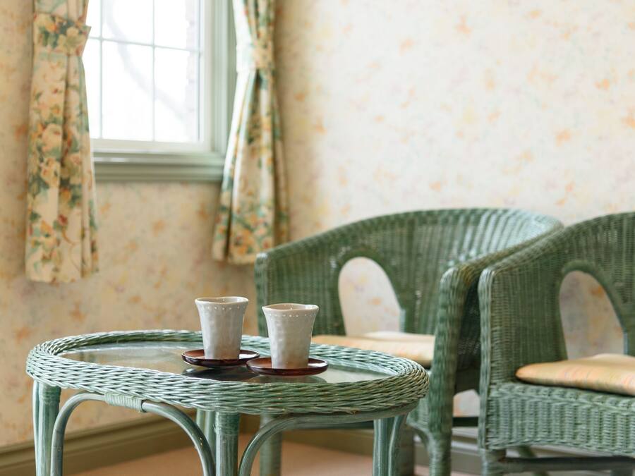 A room with a round wicker table with two white cups on coasters, a green wicker chair, and floral wallpaper.