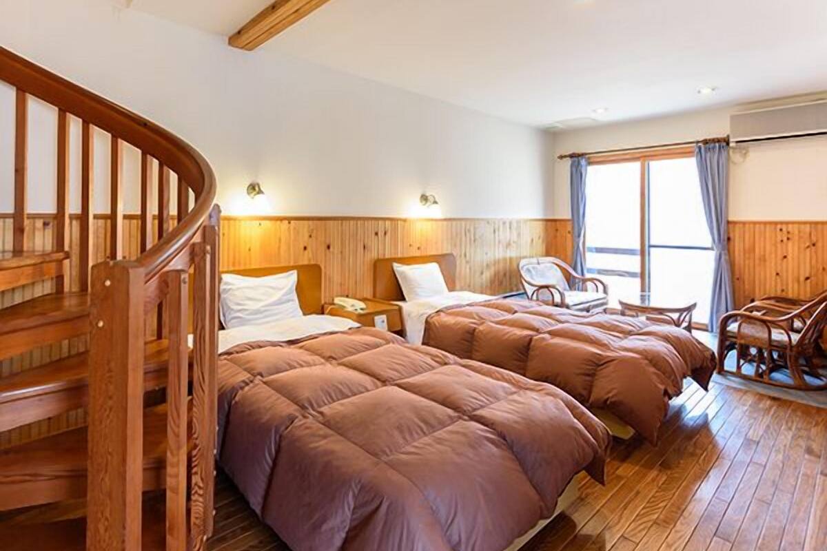 Habitación de hotel con escalera de madera, dos camas con ropa de cama marrón, cabecero de madera, suelo de madera y una ventana con cortinas.