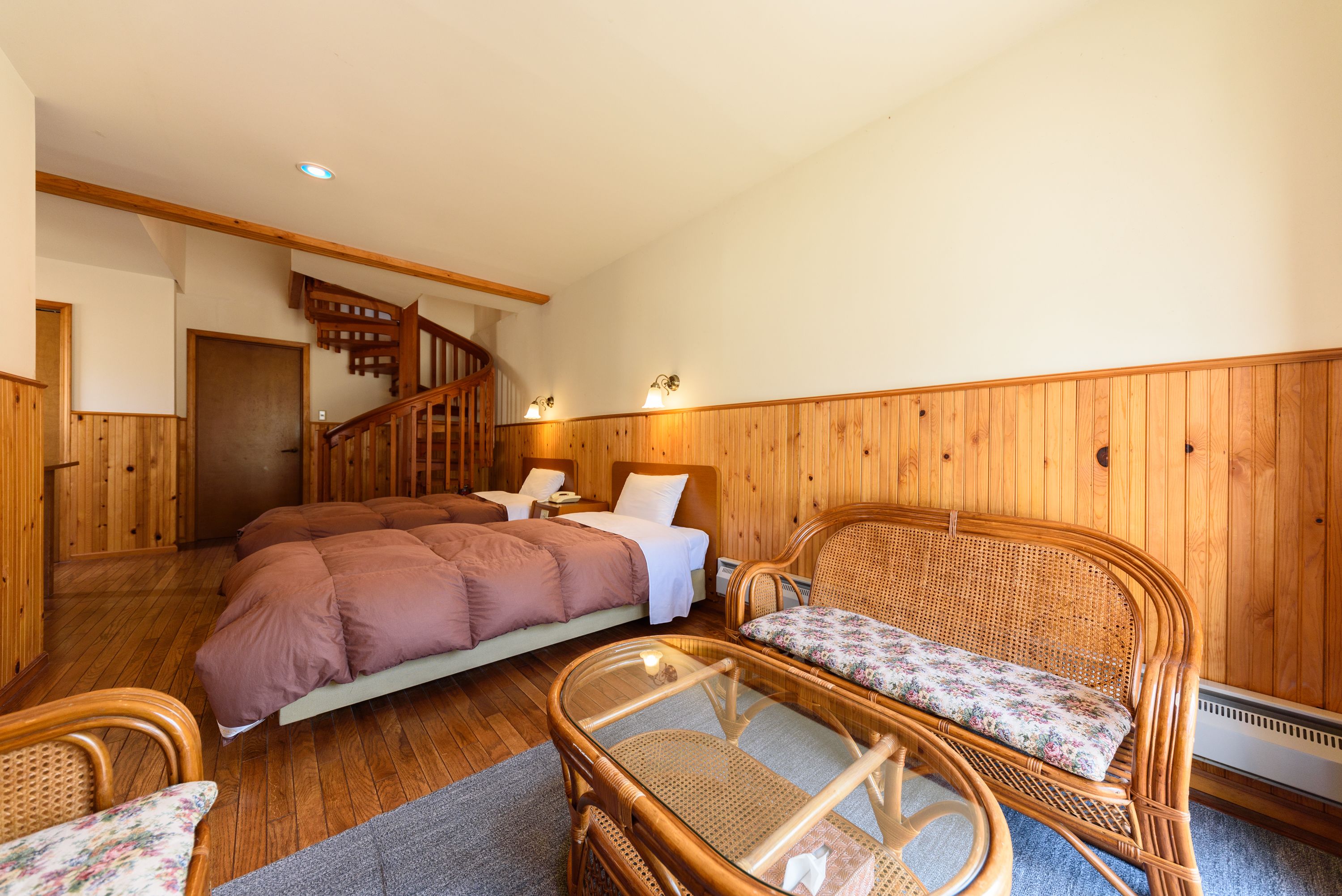 A cozy room with a wooden floor, a bed with a brown duvet, a wicker chair with a floral cushion, and a glass table.