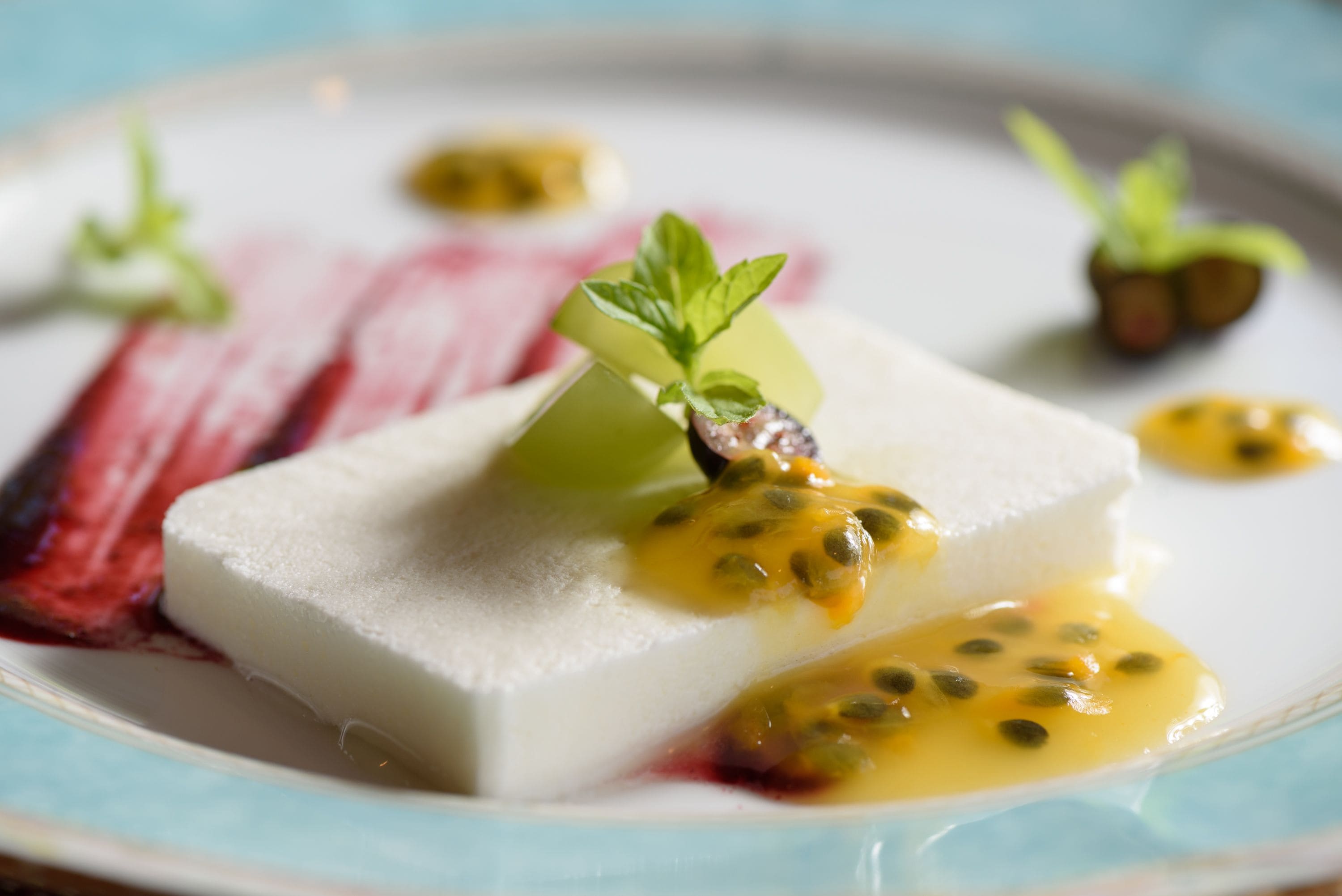 A dessert plate with a white square dessert, a green leaf garnish, and yellow sauce with seeds.
