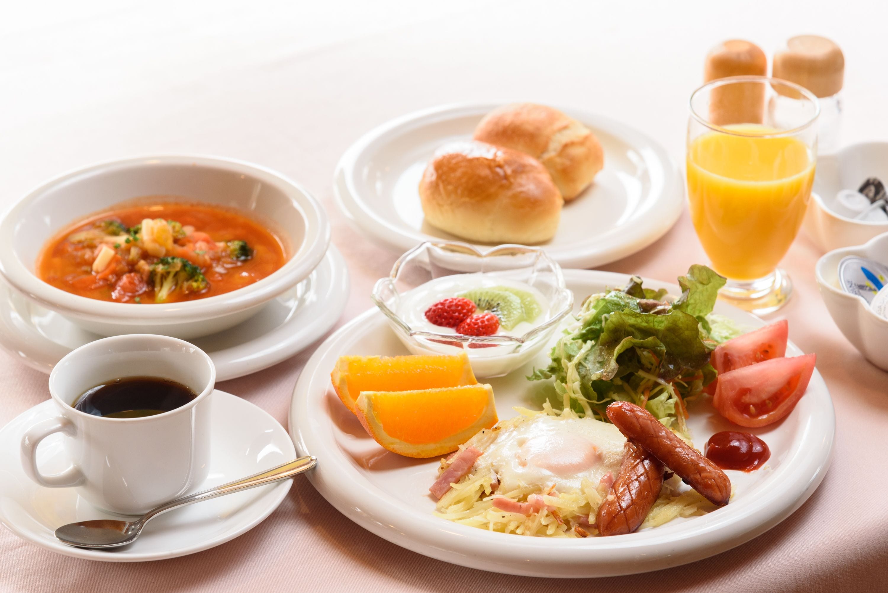 A breakfast setting with a bowl of soup, a plate with eggs, sausages, and vegetables, a glass of orange juice, and a cup of coffee.