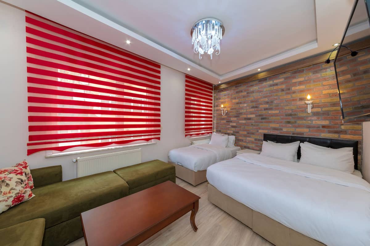 A hotel room with a brick wall, two beds, a green sofa, a wooden table, and red-striped blinds.