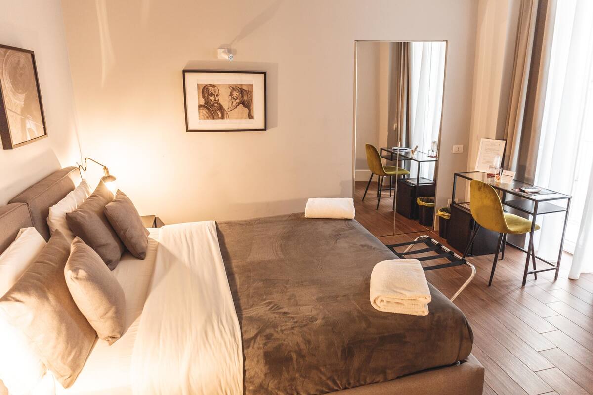 Ett hotellrum med en stor säng, ett skrivbord och en stol. På väggen hänger en inramad tavla och på den motsatta väggen finns en spegel.