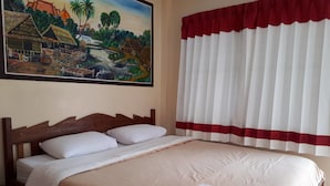 Una habitación de hotel con una cama, un cuadro de una escena de pueblo y una ventana con cortinas blancas y un ribete rojo.