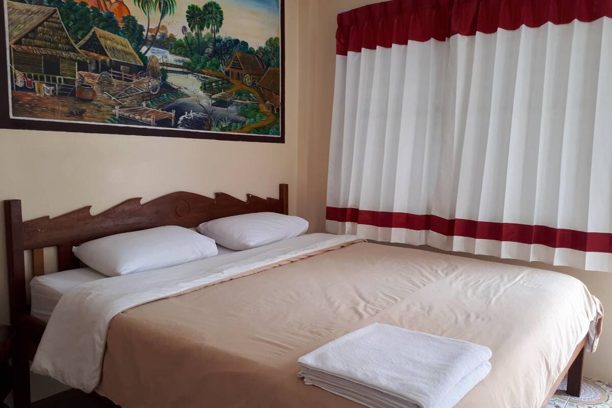 A hotel room with a bed, a painting of a village scene, and a window with white curtains and a red trim.