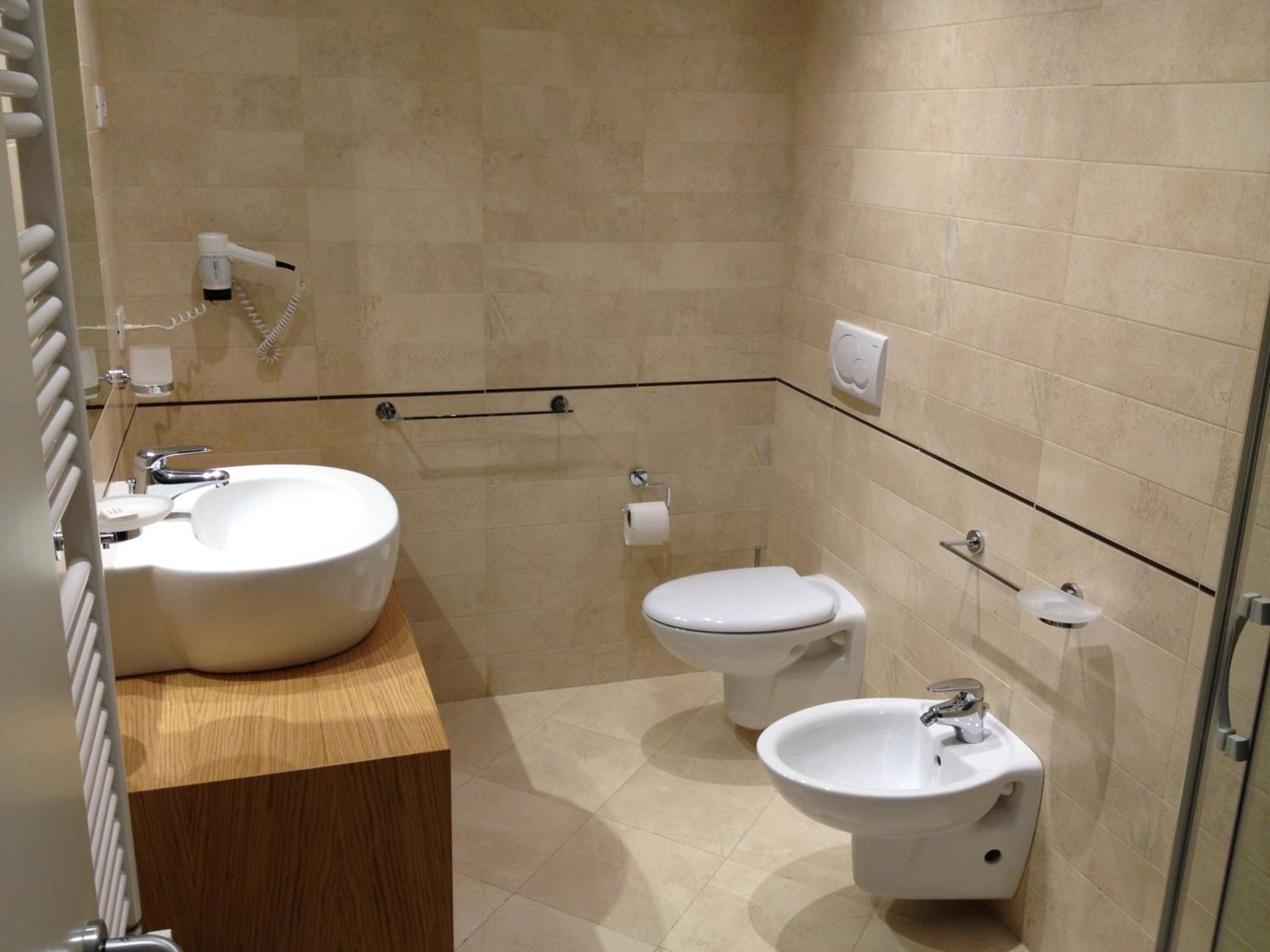A modern bathroom with a large sink, toilet, and bidet, featuring a wooden vanity and beige tiled walls.