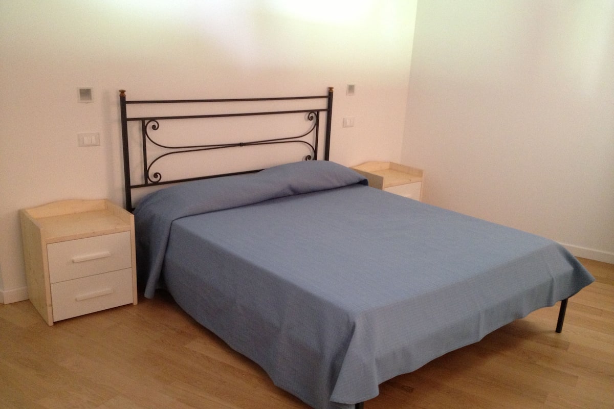 Una cama individual con estructura metálica y colcha azul en una habitación con piso de madera y paredes blancas.