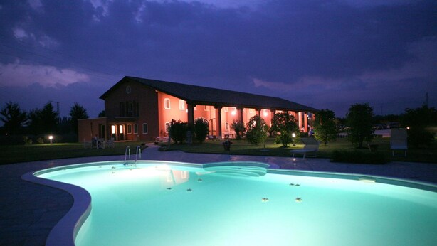 Ein Pool mit gewölbter Kante, ein Gebäude mit rotem Dach und mehreren Fenstern sowie eine gut beleuchtete Rasenfläche.