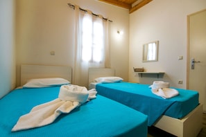 Habitación con dos camas, cada una con cubrecama azul y almohadas blancas. Hay una ventana con cortinas, un espejo en la pared y una estantería con una cesta.