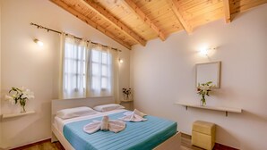 A bedroom with a bed, wooden ceiling, white walls, a window with curtains, a bedside table with a vase, and a small stool. - Athousakis Village (Chania)