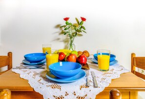 Una mesa de comedor con fuentes azules, platos y un mantel de encaje. Hay frutas, un jarrón con flores y vasos de jugo de naranja.