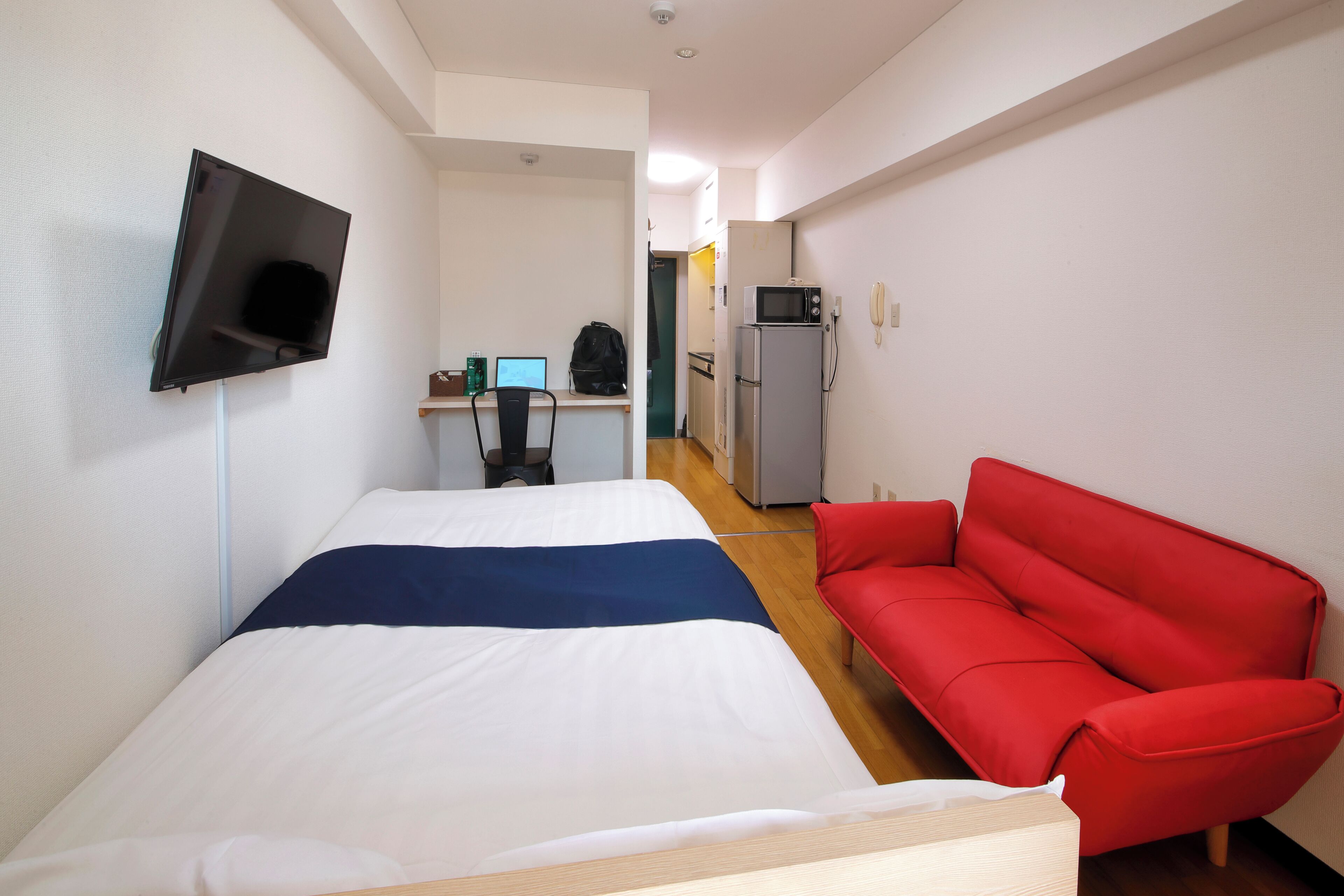 A hotel room with a bed, a red sofa, a TV mounted on the wall, a desk with a computer, and a microwave.