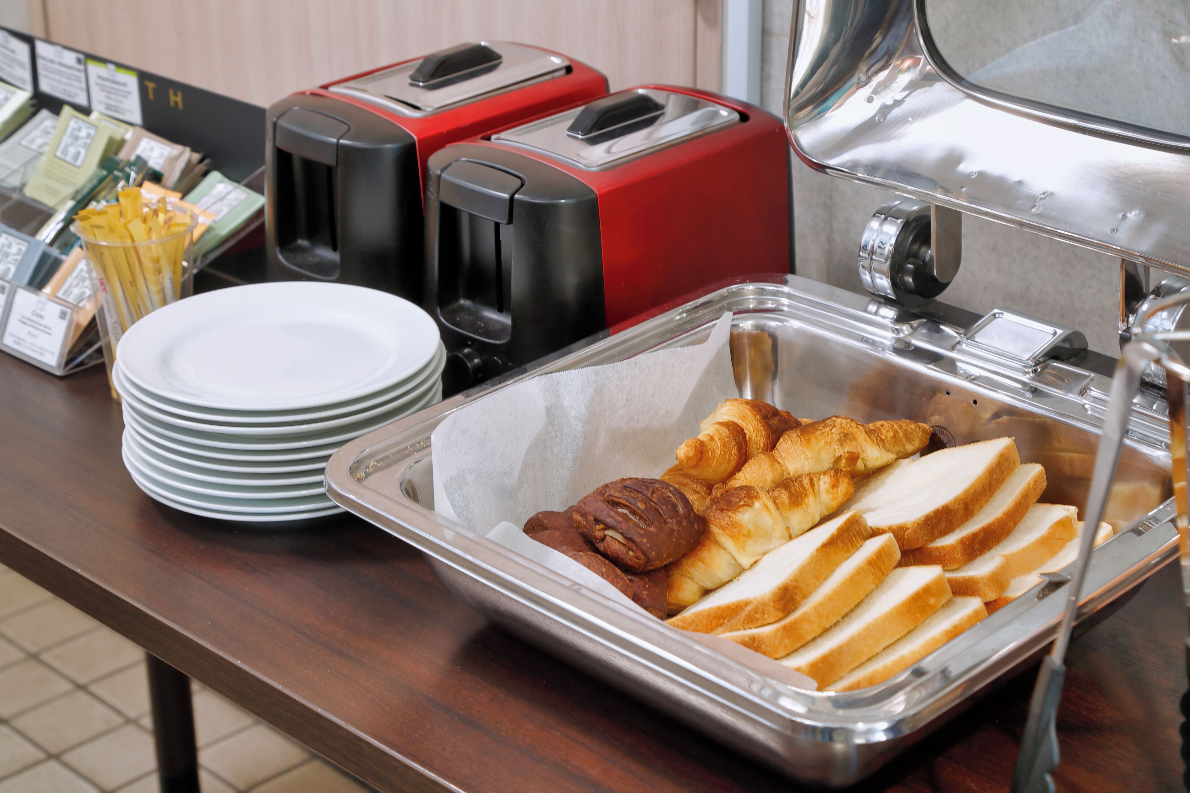 A buffet setup with a tray of bread, croissants, and a container of coffee. There are also two red toasters and a stack of white plates on the counter.