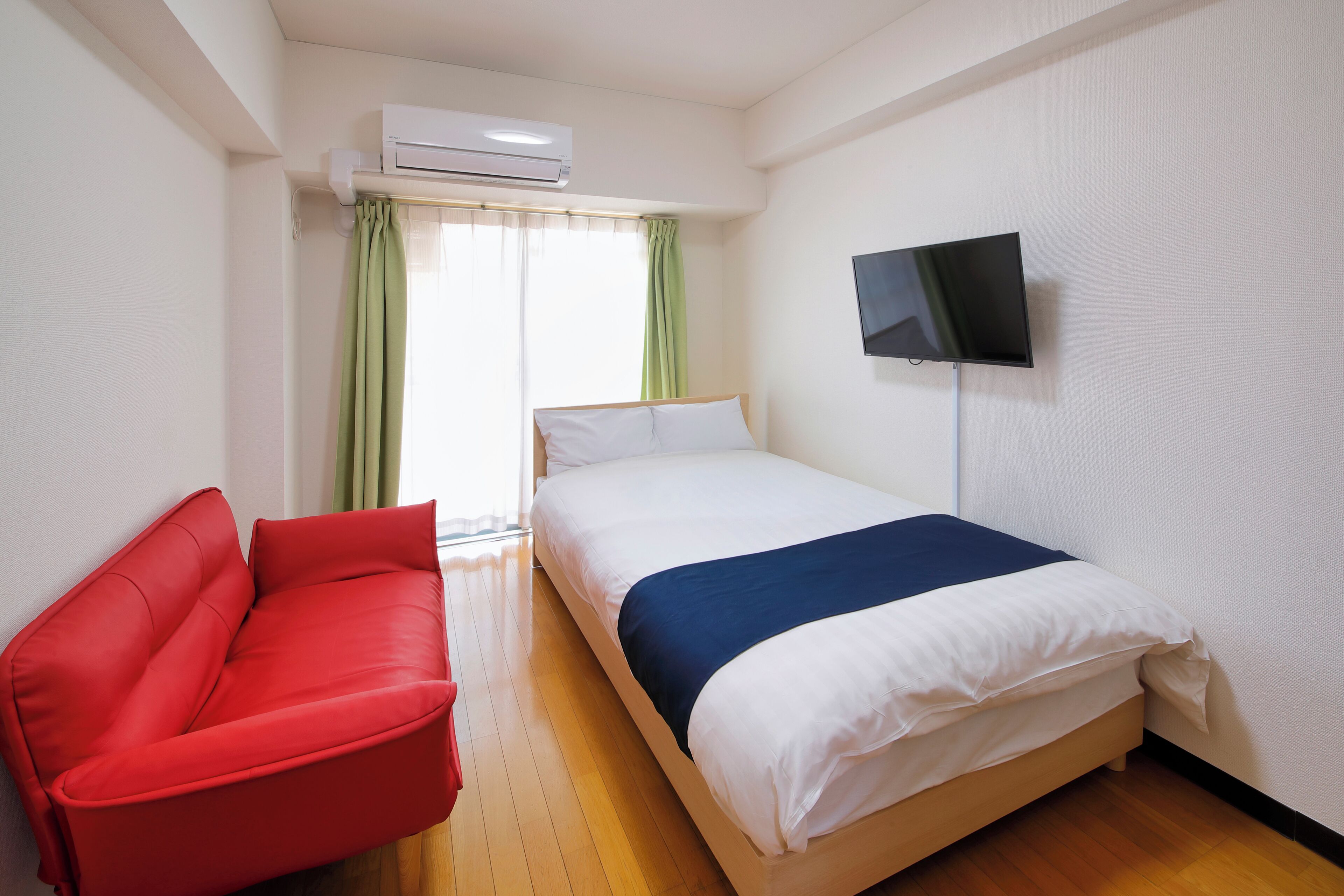 A hotel room with a bed, a red sofa, a TV on the wall, and an air conditioning unit.
