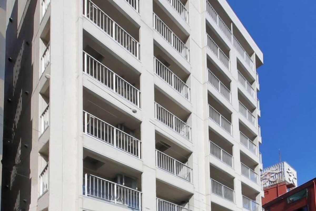 Un edificio de varios pisos con balcones, un cielo azul despejado y una vista parcial de un edificio rojo al fondo.