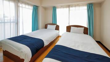 A hotel room with two single beds, wooden flooring, and large windows with curtains.