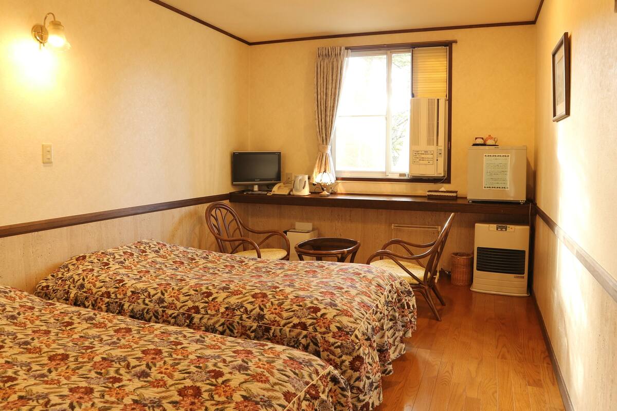 A hotel room with two beds, a desk with a computer, a window with curtains, a radiator, and a refrigerator.