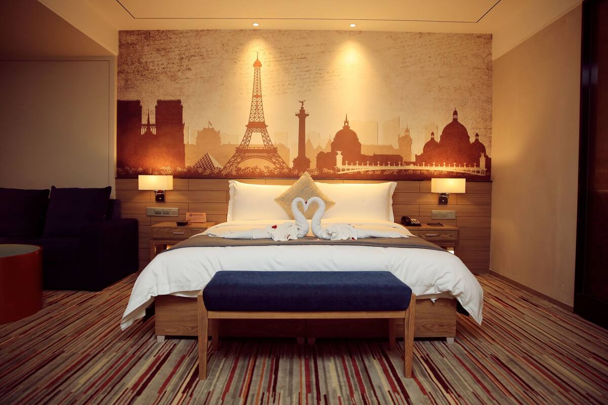 Habitación de hotel con cama, mesitas de noche, un banco, un objeto cilíndrico rojo y una pared con un mural de un paisaje urbano.