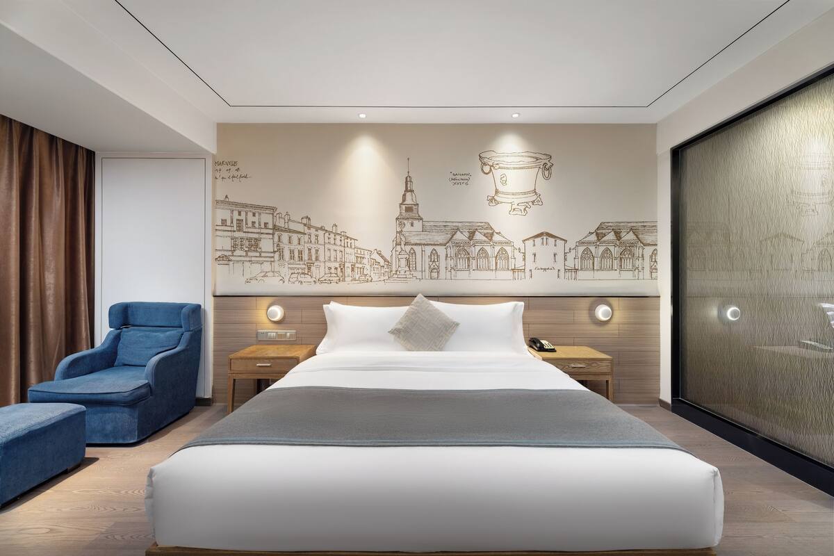 Una habitación de hotel moderna con una cama grande, un sillón azul, una mesita de noche con una lámpara y un mural en la pared que representa un paisaje urbano.