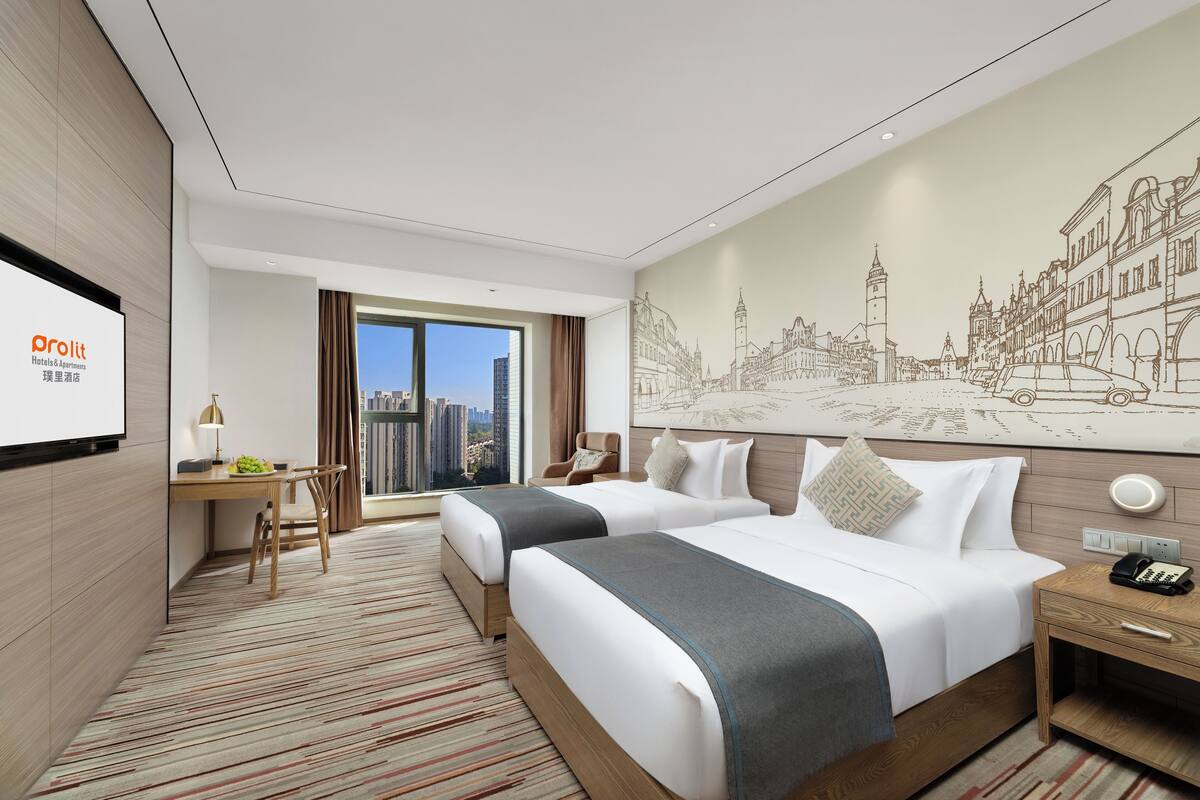 Habitación de hotel con dos camas, un escritorio, una silla, un ventanal con vistas a la ciudad y un mural en la pared.