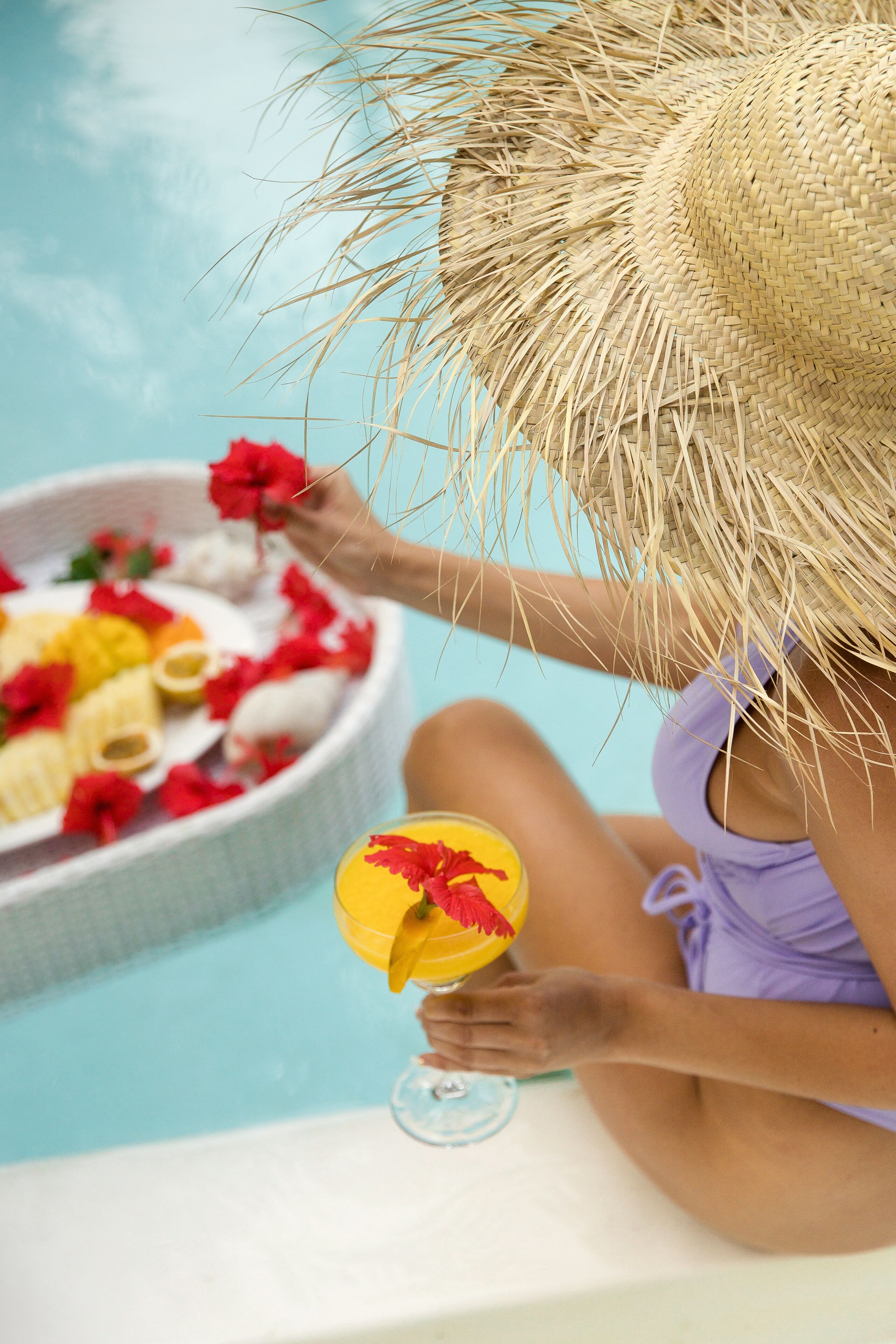 A person sitting by a pool with a straw hat, holding a drink with a straw.