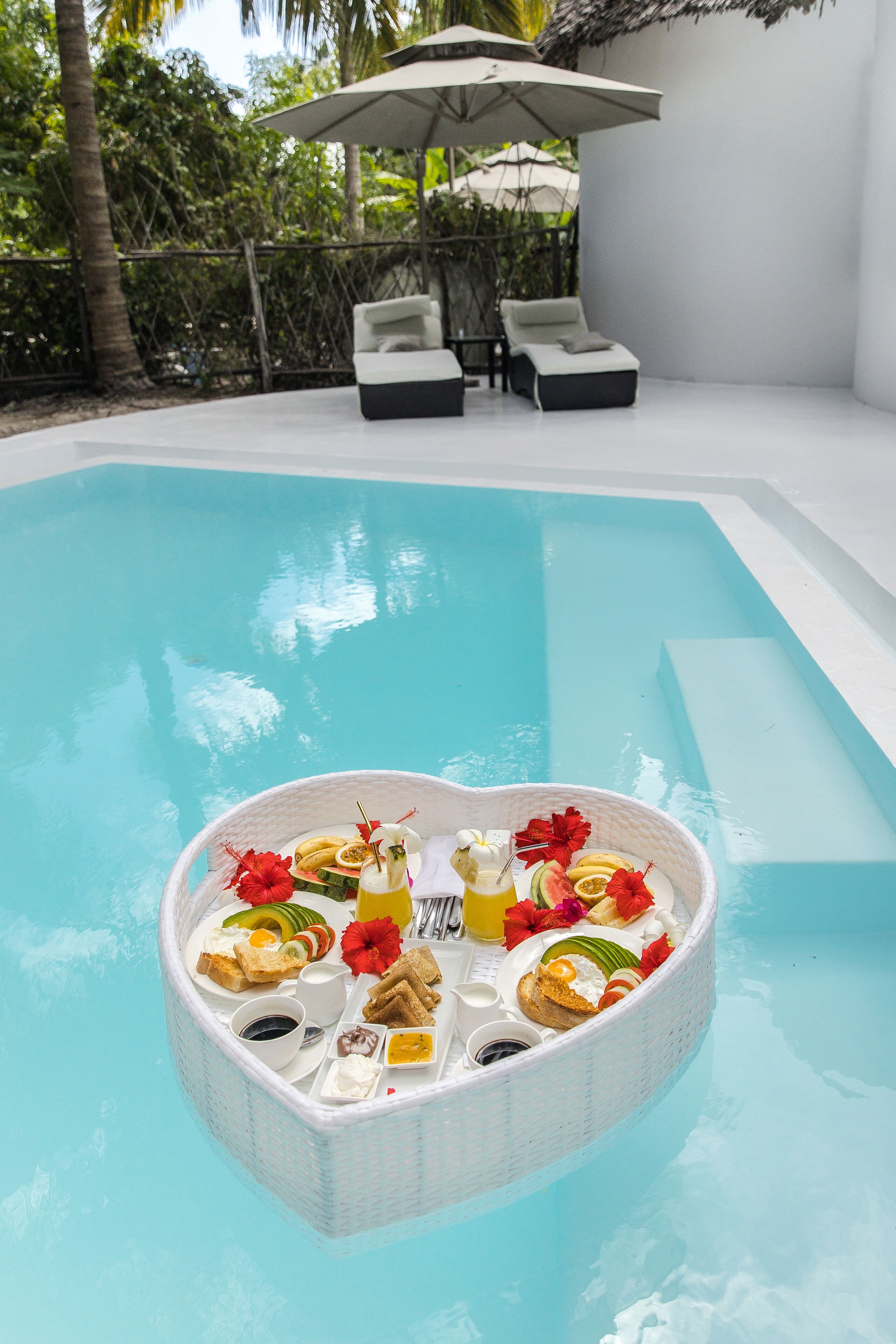 A poolside breakfast setup with a heart-shaped tray containing various foods and drinks, surrounded by lounge chairs and a palm tree in the background.