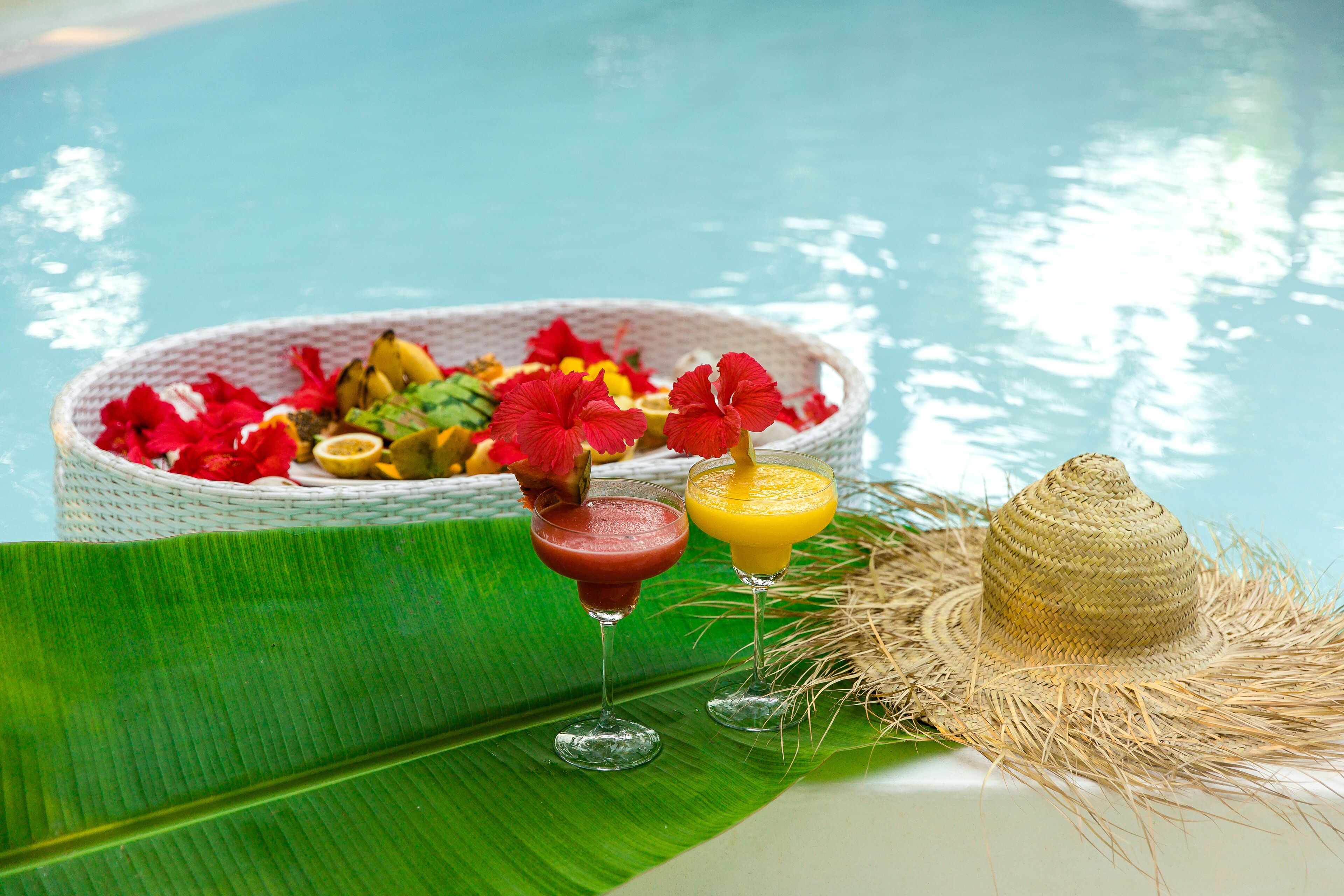 A poolside setting with a woven basket of tropical fruits and flowers, two drinks in glasses, and a straw hat on a green leaf.