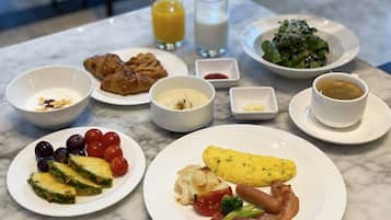 A breakfast table with plates of food, including eggs, bacon, tomatoes, and a salad. There are also bowls of yogurt and croissants, a glass of orange juice, and a cup of coffee.