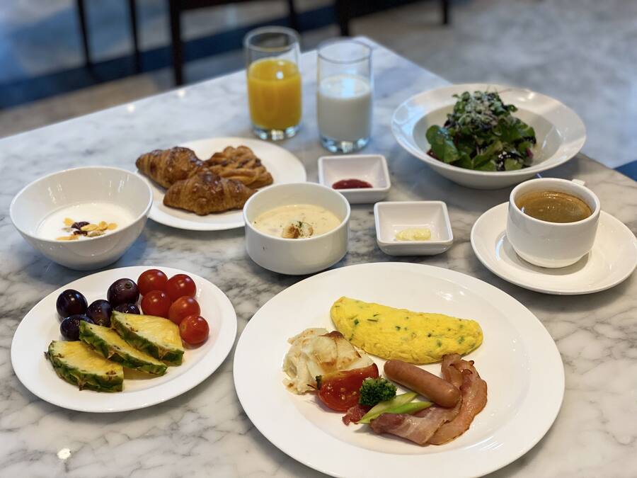 A breakfast table with plates of food, including eggs, bacon, tomatoes, and a salad. There are also bowls of yogurt and croissants, a glass of orange juice, and a cup of coffee.