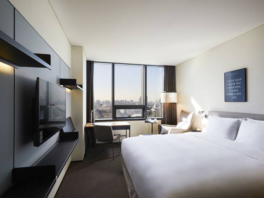 A hotel room with a bed, desk, chair, TV, and a large window with a city view.