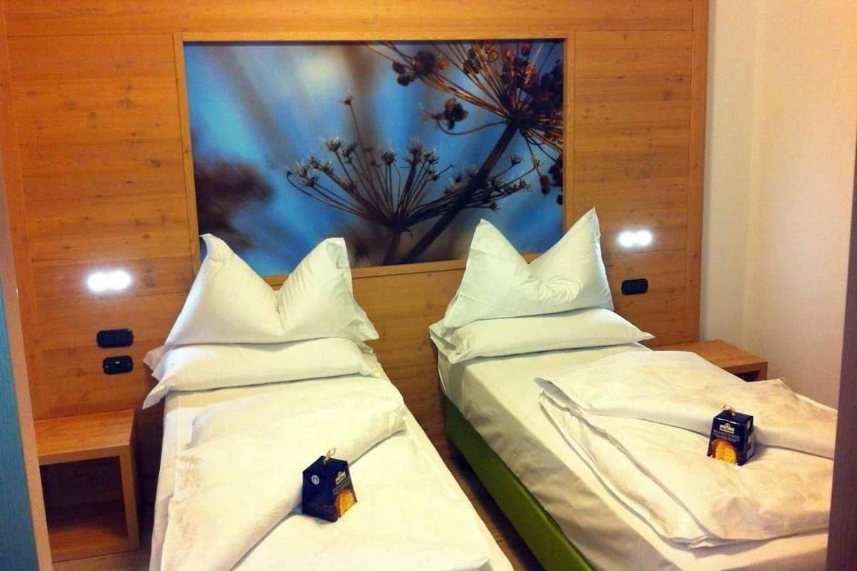 Habitación de hotel con dos camas individuales, paneles de cabecera de madera, un cuadro sobre las camas y una mesita con una caja sobre una de las camas.