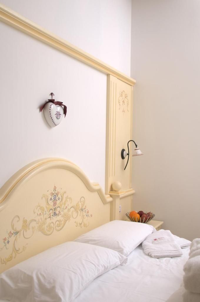 A bed with white linens, a decorative headboard, a wall-mounted light fixture, and a small bedside table with a fruit bowl.