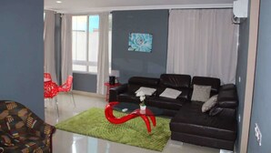 A modern living room with a dark brown sofa, a red coffee table, and a green rug. There is a window with curtains, a red chair, and a painting on the wall. - James East Legon Piece (Accra)