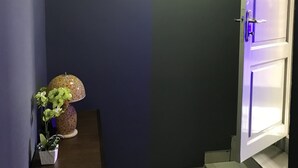 A hallway with a dark wall, a light fixture, a door with a purple light, and a small table with a lamp and a plant. - James East Legon Piece (Accra)