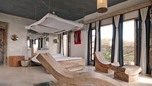 Une chambre avec un grand lit, un fauteuil en bois, une petite table et un baldaquin suspendu.