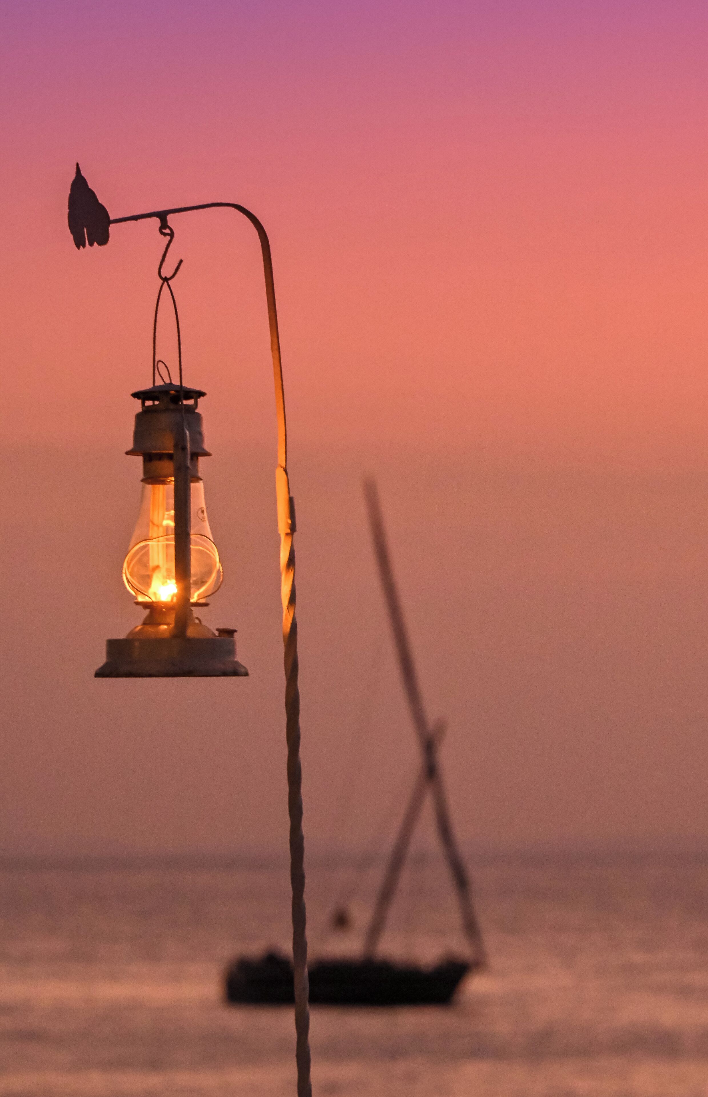 Een lantaarn met een gebogen arm, een boot op de achtergrond en een zonsondergang boven het water.