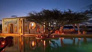 Un bâtiment traditionnel, doté d’arcades et de motifs complexes, une piscine qui reflète son architecture, et un arbre qui offre de l’ombre.